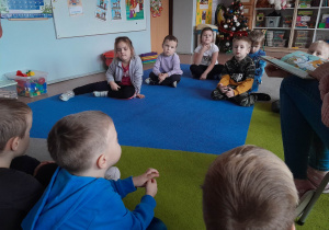 Dzieci siedzą na dywanie i słuchają opowiadania czytanego przez nauczyciela