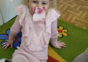 Dziewczynka pozuje do zdjęcia. Prezentuje przyłożone do twarzy zdjęcie przedstawiające opadnięty kącik ust - jeden z objawów udaru.