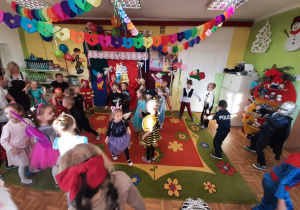 Dzieci tańczące w rytm karnawałowej muzyki. Sala przedszkolna jest kolorowa, udekorowana girlandami i serpentynami.