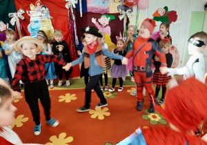 Grupa przedszkolaków tańczy trzymając się za ręce, w tle dekoracja karnawałowa.