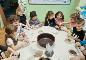 Dzieci siedzą przy stoliku, mieszają w miseczkach ziarna owsa, pszenicy, siemienia, słonecznika - kolejne ujęcie.
