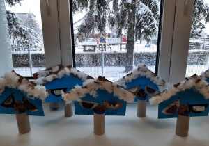 Wystawa prac plastycznych - dekoracyjne karmniki z papieru i tektury stoją na parapecie. w tle za oknami zimowy krajobraz.