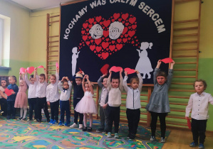 Dzieci trzymają nad głowami czerwone papierowe serca prezentując piosenkę