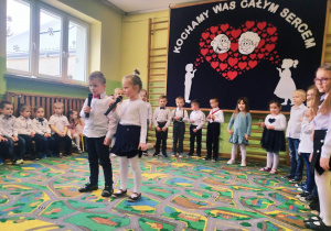 Dwoje dzieci stoi na środku i recytuje wiersz, z z tyłu dzieci stoją w półkolu