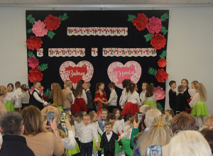 Sala OSP Wartkowice. Dzieci stoją na scenie na tle dekoracji przedstawiającej dwa duże serca, na bokach dekoracji przypięte są duże różowe i czerwone kwiaty.