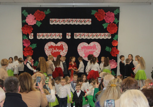 Sala OSP Wartkowice. Dzieci stoją na scenie na tle dekoracji przedstawiającej dwa duże serca, na bokach dekoracji przypięte są duże różowe i czerwone kwiaty.