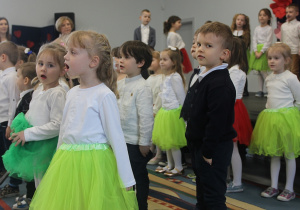 Dzieci z grupy 3,4,5 latków stojąc w rzędach śpiewają piosenkę.