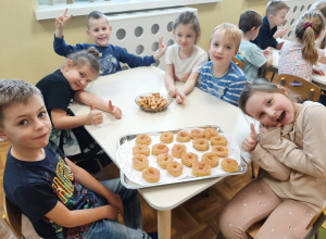 Grupa dzieci siedzi przy stoliku. Pozują pokazując tace z tłustoczwartkowymi pączkami wiedeńskimi.