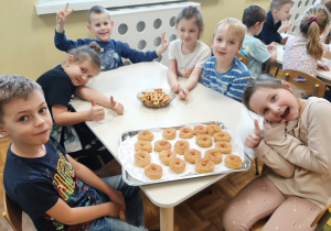 Grupa dzieci siedzi przy stoliku. Pozują pokazując tace z tłustoczwartkowymi pączkami wiedeńskimi.