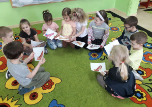Dzieci na dywanie opowiadają o wylosowanych ilustracjach pantofelka.