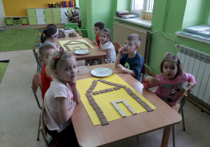 Grupa dzieci przy stoliku układa z ciasteczek piernikową chatkę Baby Jagi.
