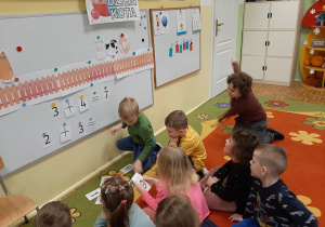 Dzieci siedzą przed tablicą na dywanie. Tworzą działania matematyczne zgodne z ilustracją na tablicy.