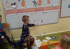 Dzieci siedzą przed tablicą na dywanie. Tworzą działania matematyczne zgodne z ilustracją na tablicy, kolejne ujęcie.