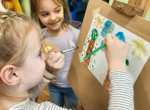 Dwie dziewczynki stoją przed sztalugą, malują farbami obrazek.