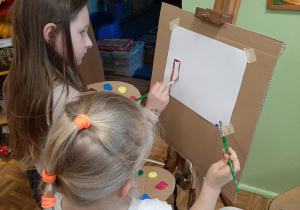 Dwie dziewczynki stoją przed sztalugą, malują farbami obrazek.