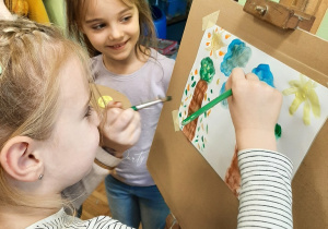Kolejne dwie dziewczynki stoją przed sztalugą, malują farbami obrazek.