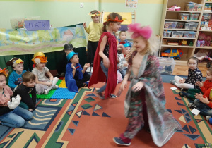 Dzieci bawią się w teatr. Na scenie w sali przedszkolnej tańczą dwie dziewczynki. w tle scena ze scenografią przedstawiającą las.