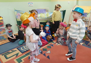 Dzieci bawią się w teatr. Na pierwszym planie dziewczynka w czapce lisa i chłopiec w niebieskim kapeluszu. W tle scena, dekoracja.