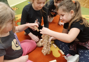 Dziewczynki na dywanie konstruują budowlę z klocków drewnianych.