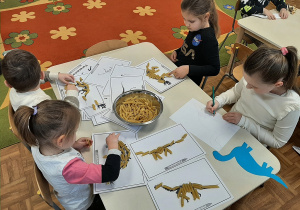 Dzieci siedzą przy stoliku, układają według wzoru szkielety dinozaurów, używając makaronu
