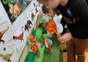 Przedszkolaki tworzą makietę prahistorycznej krainy. Umieszczają papierowe drzewa, makietę wulkanu i figurki dinozaurów.