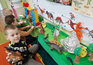 Grupa dzieci bawi się figurkami dinozaurów na przygotowanej makiecie. W tle gazetka tematyczna.