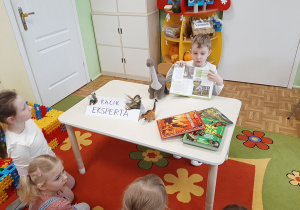 Przy stoliku siedzi kolejne dziecko i opowiada pozostałym siedzącym naprzeciw na dywanie o swoim ulubionym dinozaurze.