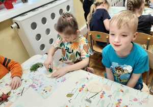 Dzieci siedzą przy stolikach odciskają w masie solnej ślady dinozaurów - figurek i roślinności, kolejne ujęcie.
