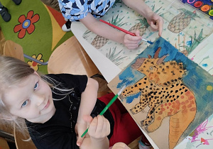 Dzieci przy stoliku malują farbami obrazki dinozaurów.