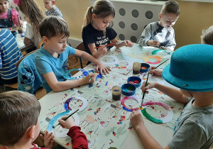 Dzieci siedzą pry stoliku, malują farbami szablon cyfry 8.