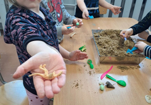 Dzieci stoją przy stoliku i szukają szkieletów dinozaurów ukrytych w piasku