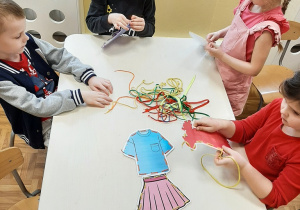 Grupa dzieci przy stoliku przewlwka kolorowe sznurki przez otwory w szblonach.