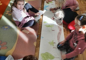 Dzieci na podłodze na długim pasie kartonu tworzą ilustrację - tło do bajki o czerwonym kapturku.