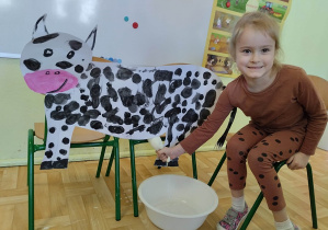 Dziewczynka pozująca do zdjęcia przy tekturowym szablonie krowy. Bierze udział w zabawie "Doimy krowę".