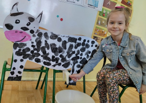 Kolejna dziewczynka pozująca do zdjęcia przy tekturowym szablonie krowy. Bierze udział w zabawie "Doimy krowę".