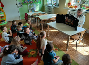 Dzieci siedzą na dywanie, pani bibliotekarka czyta bajkę "Słowik" korzystając z teatrzyku kamishibai.