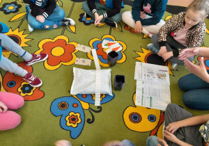 Dzieci z grupy 5-6 latków siedzą na dywanie i oglądają zawartość pudełka