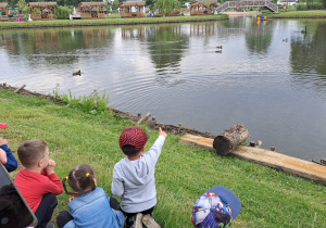 Dzieci siedzą w pobliżu stawu i oglądają kaczki i łabędzie.
