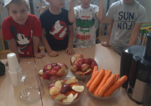 Przedszkolaki stoją w czapkach kucharskich przed obranymi warzywami i owocami