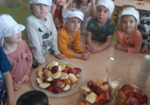 Przedszkolaki stoją w czapkach kucharskich przed obranymi warzywami i owocami