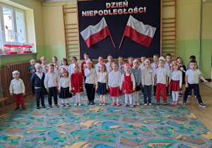 Dzieci na holu przedszkolnym śpiewają hymn Polski. W tle dekoracja z napisem Święto Niepodległości.