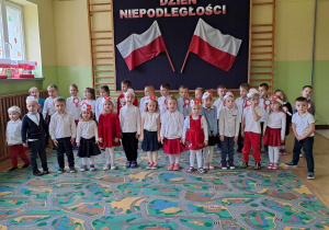 Dzieci na holu przedszkolnym śpiewają hymn Polski. W tle dekoracja z napisem Święto Niepodległości.
