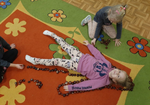 Dziewczynka leżąca na dywanie obłożona wokół kasztanami.Przy niej koleżanki
