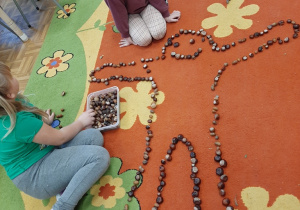 Dzieci bawią się na dywanie kasztanami. Układają sylwetkę człowieka.