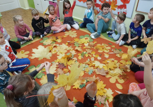 Grupa dzieci siedzi na dywanie w kole. na środku dywanu leżą jesienne liście.