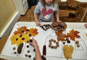 Dzieci przy stoliku tworzą na dużym szablonie jesienną kompozycję domu i podwórka z materiału przyrodniczego