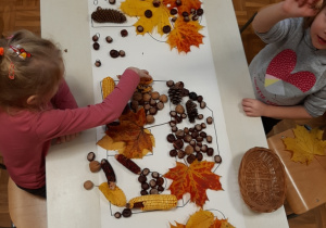 Dzieci przy stoliku tworzą na dużym szablonie jesienną kompozycję domu i podwórka z materiału przyrodniczego, kolejne ujęcie.