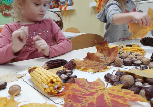Dziewczynki przy stole bawią się jesiennymi skarbami: kasztanami, liśćmi, kolbami kukurydzy.