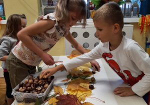 Dzieci przy stoliku tworzą na dużym szablonie jesienną kompozycję krokodyla z materiału przyrodniczego