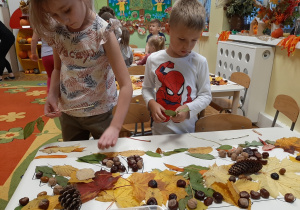 Dzieci przy stoliku tworzą na dużym szablonie jesienną kompozycję krokodyla z materiału przyrodniczego, inne ujęcie.